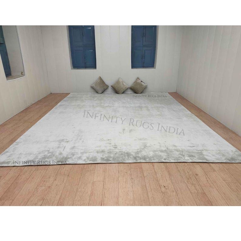 Puede incluir: Una alfombra rectangular blanca con un patr&oacute;n sutil y desgastado. La alfombra est&aacute; colocada sobre un suelo de madera en una habitaci&oacute;n con dos ventanas y tres almohadas. El texto "INFINITY RUGS INDIA" est&aacute; impreso en la alfombra.