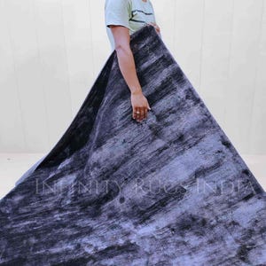 May include: A person holding a large, dark blue and gray rug with a subtle, textured pattern. The rug has a faded, vintage look and the text "INFINITY RUGS INDIA" is visible in the lower center of the rug.