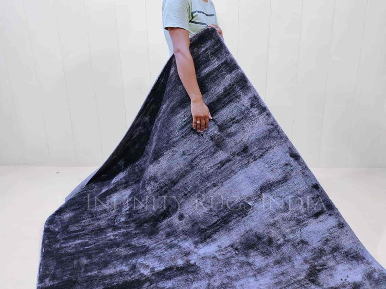 May include: A person holding a large, dark blue and gray rug with a subtle, textured pattern. The rug has a faded, vintage look and the text "INFINITY RUGS INDIA" is visible in the lower center of the rug.