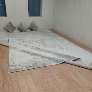 Puede incluir: Una alfombra blanca y esponjosa con un sutil patr&oacute;n gris. La alfombra est&aacute; doblada por la mitad y colocada sobre un suelo de madera. La alfombra est&aacute; rodeada por tres cojines beige.