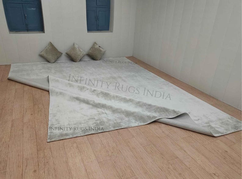 Puede incluir: Una alfombra blanca y esponjosa con un sutil patr&oacute;n gris. La alfombra est&aacute; doblada por la mitad y colocada sobre un suelo de madera. La alfombra est&aacute; rodeada por tres cojines beige.