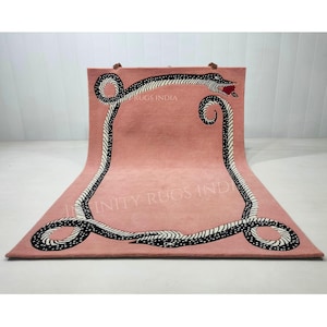 Puede incluir: Una alfombra rosa con un diseño de serpiente en blanco y negro. La serpiente está enroscada alrededor del borde de la alfombra. El texto "RUGS INDIA" e "INFINITY RUGS IND" es visible en la alfombra.