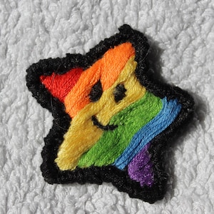 Könnte beinhalten: Ein gestickter Stern in Regenbogenfarben mit einem lächelnden Gesicht. Der Stern ist schwarz mit einem Regenbogenmuster und einem violetten Akzent.
