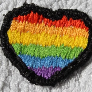 Könnte beinhalten: Ein regenbogenfarbener, herzförmiger gestickter Aufnäher mit schwarzem Rand. Die Regenbogenfarben sind rot, orange, gelb, grün, blau und lila.