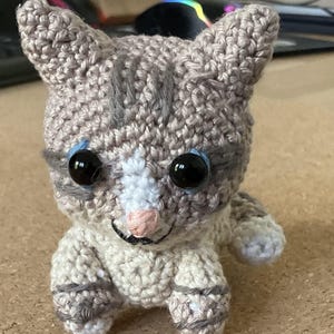 Puede incluir: Un gato de crochet gris y blanco con ojos azules. El gato está sentado sobre una superficie marrón.