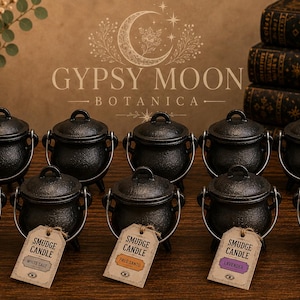 May include: Ten black cast iron cauldron-shaped candles with lids and metal handles arranged in two rows. Each front-row cauldron features a beige paper tag labeled SMUDGE CANDLE with a colored square indicating the scent: rose, white sage, palo santo, lavender, or sandalwood.