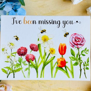 May include: A white card with the text "I've been missing you" in black cursive writing. The card is decorated with watercolor illustrations of flowers, including white daffodils, red tulips, pink roses, and white daisies. There are also small yellow and black bees flying around the flowers.