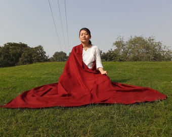 May include: A white long-sleeved shirt with a textured pattern paired with a large, solid deep red wrap or shawl that drapes over the shoulders and extends across the ground in a wide, flowing fabric display.