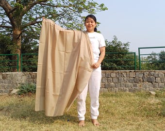 May include: A person is holding a light brown shawl in front of a green fence and a stone wall. The shawl is made of a soft, lightweight fabric and has a subtle sheen.
