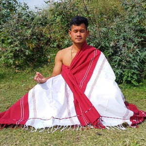 Peut inclure: Une personne est assise en tailleur sur l'herbe, vêtue d'un châle rayé blanc et rouge avec des glands. Le châle est drapé sur les épaules de la personne et noué à la taille.