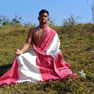 Peut inclure: Une personne est assise en tailleur sur une colline herbeuse portant un dhoti en coton blanc et rose. La personne porte un collier de perles autour du cou.