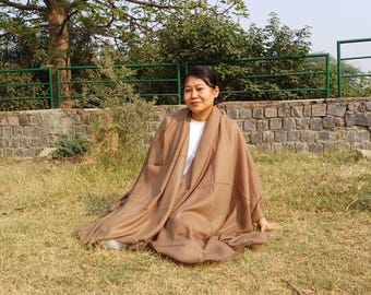 May include: A woman is sitting on the ground wearing a brown shawl. She is sitting in a grassy area with a stone wall behind her.