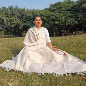 May include: A person sits cross-legged on the grass, wrapped in a cream-colored throw blanket with white fringe. The blanket has a subtle striped pattern.