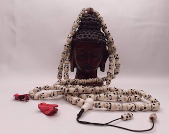 May include: A white bone mala with skull beads, a black cord, and a white bone skull bead tassel. The mala is draped over a wooden statue of a Buddha head.