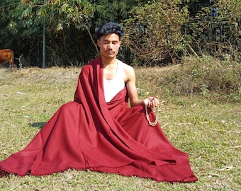 May include: A person in a red robe sits cross-legged on grass, eyes closed, holding a beaded necklace. They wear a white undershirt. A cow grazes in the background. The scene is outdoors on a sunny day.