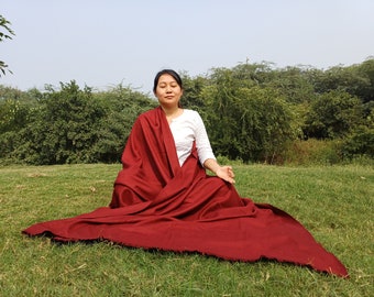 May include: A woman sits cross-legged on a green grassy field, wearing a deep red shawl draped over a white shirt. She has her eyes closed and her hands are in a meditative position.