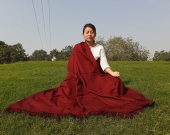May include: A large, deep maroon rectangular shawl with a textured weave and fringed edges. The shawl is draped over the shoulders and lap, featuring a consistent solid color throughout the fabric and a visible fringe detail along the bottom hem.