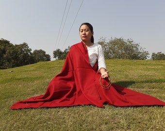 May include: A person seated on a grassy hill, draped in a vibrant red shawl. The shawl is spread out, creating a wide, flowing shape. The person is wearing a white top and holding gold bangles. The scene is set outdoors on a sunny day.