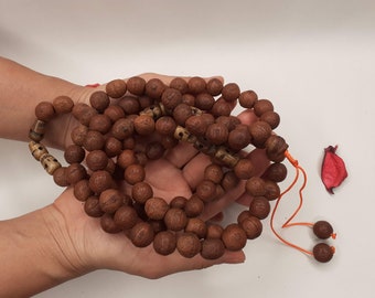May include: A brown wooden mala bead necklace with a knotted tassel. The beads are round and have a textured surface. The necklace is held in a person's hands.