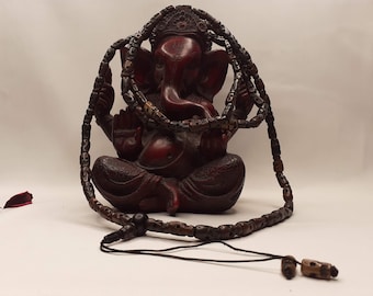 May include: A dark brown wooden beaded necklace with a black bead accent. The necklace is wrapped around a red wooden statue of the Hindu deity Ganesha.