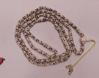 May include: A white prayer bead necklace with black markings. The beads are cylindrical and have a small hole in the center. The necklace is strung on a yellow cord.
