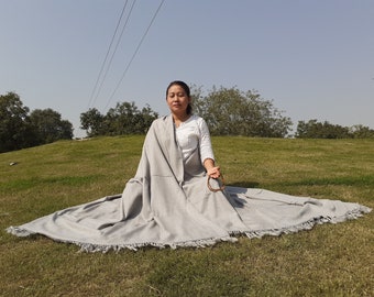 May include: A person sits cross-legged on a grassy hill wearing a gray shawl with fringe. The shawl is draped around the person's shoulders and is pulled up to their chin.