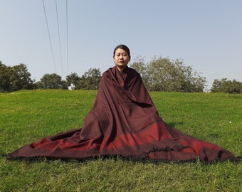 May include: A burgundy shawl with a red stripe and black fringe is spread out on a grassy field. The shawl is draped over a person sitting cross-legged with their eyes closed. The background features trees and a clear sky.