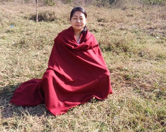 May include: A solid deep red shawl or blanket draped over the shoulders and body, featuring a smooth, opaque fabric texture with clean, finished edges that extend down to the ground in a simple, unpatterned design for warmth and coverage.