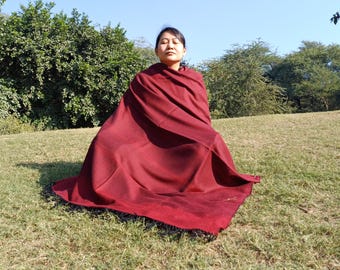 May include: A large, solid deep maroon shawl with a smooth texture and dark fringe along the bottom edge. The fabric is draped over the shoulders and body, extending outward to cover the lap and legs in a wide, flowing, and rectangular shape.