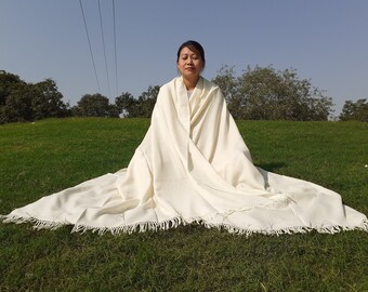 Puede incluir: Un chal de pashmina blanco con borde de flecos. El chal está drapeado sobre una persona sentada en una colina cubierta de hierba.