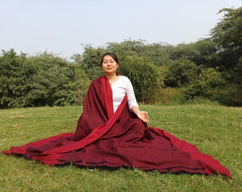 May include: A white long-sleeved shirt paired with a large, rectangular shawl featuring a deep maroon and dark burgundy color scheme. The shawl is draped over the shoulders and lap, showcasing a solid texture with a dark fringe detail at the edge.