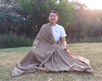 May include: A woman sits cross-legged on the grass wearing a brown shawl and a white t-shirt. She has her eyes closed and her hands resting in her lap.