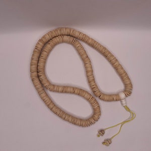 Op de afbeelding: Een beigehouten mala-ketting met een witte kraal aan het einde en een gele kwast. De ketting is gemaakt van ronde kralen en ligt op een wit oppervlak.