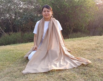 May include: A person wearing a white shirt is sitting on the grass with a large, light brown scarf draped around their shoulders. The scarf is made of a soft, flowing fabric.