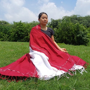 Peut inclure: Une personne est assise en tailleur sur une pelouse verte portant un châle rayé rouge et blanc avec des glands. Le châle est drapé sur les épaules de la personne et est plié en deux.