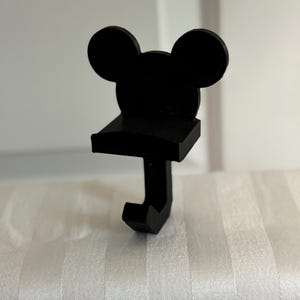 Könnte beinhalten: Ein schwarzer Haken in Form von Mickey Mouse. Der Haken hat die ikonischen Micky-Maus-Ohren oben, ein kleines Regal und einen nach unten gerichteten Haken. Der Artikel besteht aus einem mattschwarzen Material und ist zum Aufhängen von Gegenständen bestimmt.