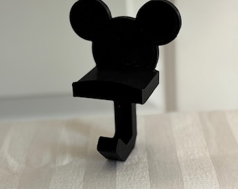 May include: A black, Mickey Mouse-shaped hook. The hook features the iconic mouse ears at the top, a small shelf, and a downward-facing hook. The item is made of a solid, matte black material and is designed to hang items.