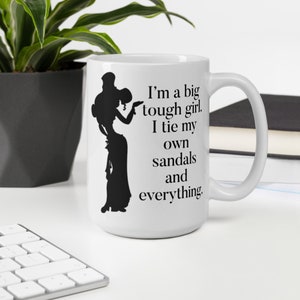 Puede incluir: Taza de cerámica blanca con una silueta negra de una mujer atándose las sandalias. La taza tiene el texto "I'm a big tough girl. I tie my own sandals and everything."