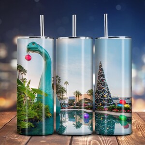 Puede incluir: Tres vasos de acero inoxidable con diferentes diseños navideños. El primer vaso presenta un dinosaurio verde sosteniendo un adorno rojo. El segundo vaso presenta un árbol de Navidad con adornos y luces. El tercer vaso presenta un árbol de Navidad con adornos y luces reflejados en una piscina.