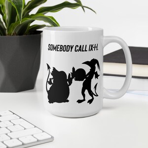 May include: White ceramic mug with a black graphic of two cartoonish figures and the text "SOMEBODY CALL IX-H." The mug has a large handle and is sitting on a white surface.