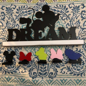 Könnte beinhalten: Schwarzes Holzschild mit dem Wort "DREAM" in großen Buchstaben. Eine schwarze Silhouette von Mickey Mouse befindet sich über dem Wort. Fünf kleine Figuren von Mickey Mouse-Charakteren befinden sich unter dem Schild, in Schwarz, Rot, Grün, Pink und Blau.