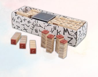 May include: A wooden alphabet stamp set with a black ink pad, presented in a box decorated with black letter patterns. Several individual stamps are displayed, showing the red rubber stamp faces. The set is ideal for crafting and scrapbooking.