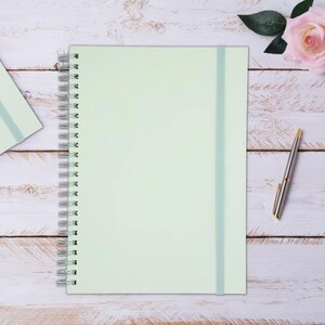 Op de afbeelding: Een lichtgroen spiraalgebonden notitieboek met een elastische bandsluiting. Een zilveren en gouden pen ligt op een wit houten oppervlak naast een roze roos. Een kleiner notitieboek is linksboven zichtbaar.