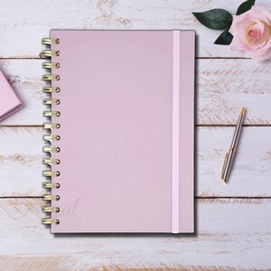 Op de afbeelding: Een lichtroze notitieboek met gouden spiraalbinding en bijpassende elastische sluiting. Een zilveren pen met gouden details ligt ernaast. Op de achtergrond ligt een roze roos. Perfect voor notities en planning.