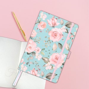 Op de afbeelding: Een notitieboek met een lichtblauwe kaft versierd met roze rozen en groene bladeren. Een gouden pen ligt naast de open, gestippelde pagina's van een tweede notitieboek. Een roze elastiek sluit het notitieboek af.