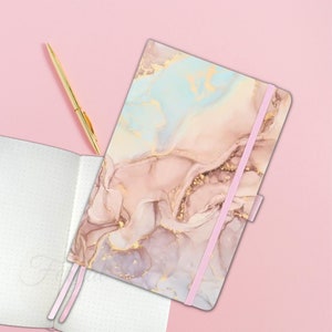 May include: A notebook with a pink elastic closure and a cover featuring a marble design in shades of pink, gold, and blue. A gold pen rests beside the notebook, and an open notebook with dotted pages is also visible.