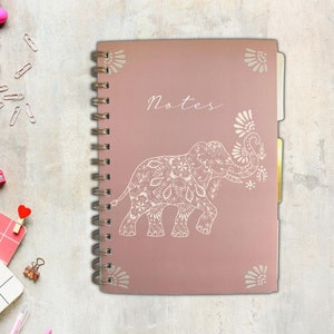 May include: A spiral-bound notebook with a dusky pink cover. The cover features a detailed white elephant design and the word "Notes" in script. Decorative floral accents are in each corner. The notebook has gold-coloured dividers.