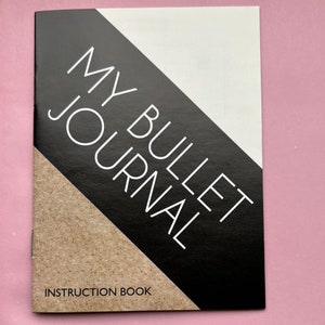 May include: A closed instruction book for a bullet journal. The cover has a black diagonal stripe with the words "MY BULLET JOURNAL" in white. The bottom of the cover reads "INSTRUCTION BOOK". The background is a light pink.