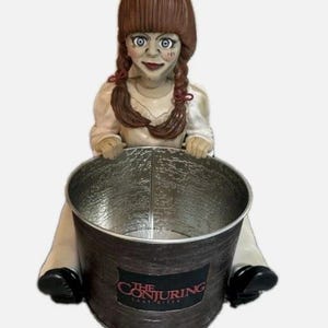 Może przedstawiać: Doniczka w kształcie lalki z brązowymi warkoczami, niebieskimi oczami i białą sukienką. Lalka trzyma metalowe wiadro z napisem "THE CONJURING: LAST RITES" z przodu. Wiadro ma ciemne, teksturowane wykończenie.