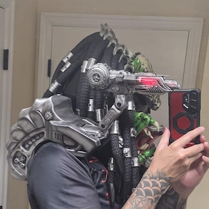 May include: A Predator-style helmet featuring long black dreadlocks with silver rings, a metallic silver shoulder cannon with a red light, and a matching silver shoulder armour piece. The helmet has green textured details and a complex mechanical design.
